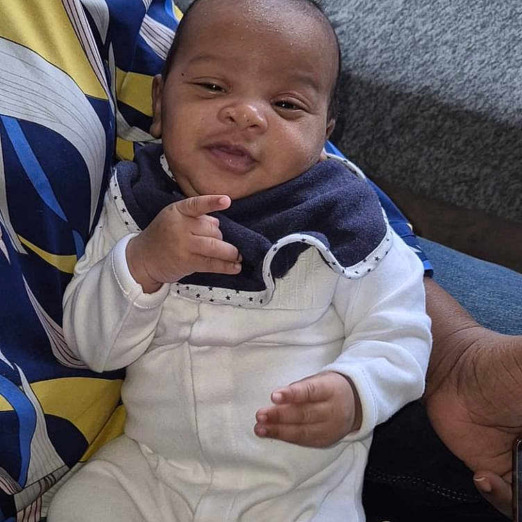 Marcus a rejoint le concours — aidez-le/la à gagner de superbes lots ! adult_hand, baby, bib, casual, closeup, comfort, couch, denim, face, hand_gesture, holding, indoor, infant, patterned_shirt, person, portrait, seated, skin, smiling, white_clothing