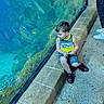 child, boy, aquarium, glass, reflection, tank_top, yellow_clothing, stone_ledge, fish, water, underwater, casual_clothing, shorts, shoes, person, curious, sitting, indoor, blue, reflection_in_glass