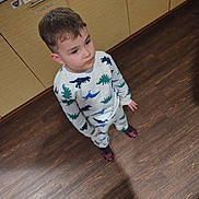 Raiden is registered to the contest to win money with this photo: child, toddler, pajamas, dinosaur_print, standing, wooden_floor, kitchen, cabinet, light_wood, curly_hair, looking_up, indoor, footwear, sneakers, casual_clothing, expression, young_child, person, home, flooring