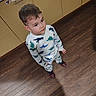 child, boy, dinosaur_pajamas, standing, wooden_floor, kitchen, cabinets, curious, indoors, clothing, footwear, short_hair, young, person, casual, expression, looking_up, home, flooring, light