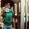 toddler, child, afro_hair, green_tshirt, nirvana_logo, bottle, crib, nursery, jeans, indoor, standing, curly_hair, baby, furniture, carpet, hand, holding, person, cute, casual_clothing