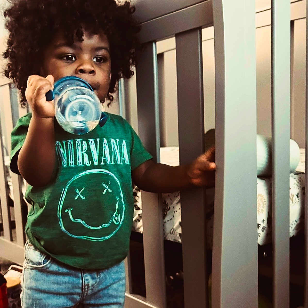 Logan joined the competition — help win amazing prizes! toddler, child, afro_hair, green_tshirt, nirvana_logo, bottle, crib, nursery, jeans, indoor, standing, curly_hair, baby, furniture, carpet, hand, holding, person, cute, casual_clothing