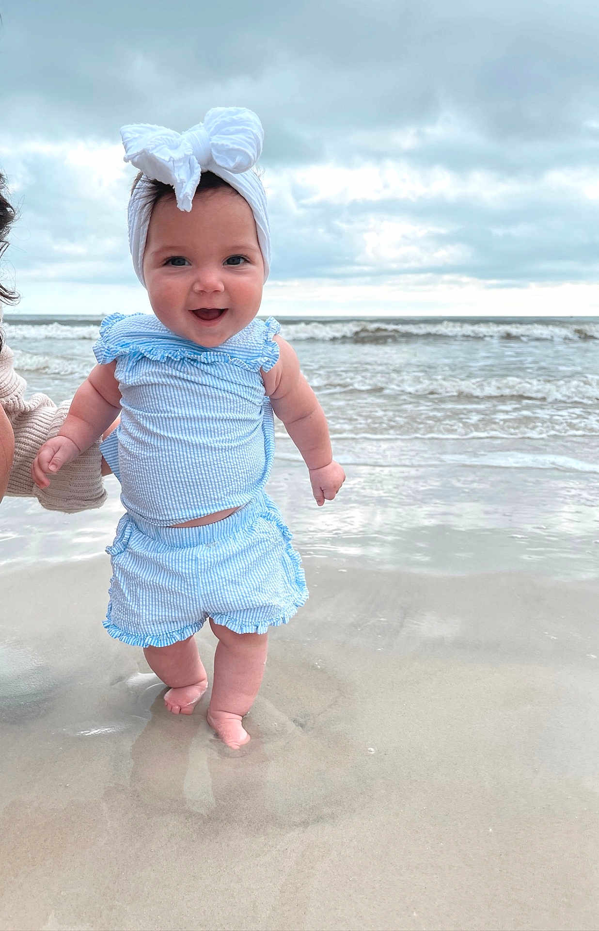 Lilah is registered to the contest to win money with this photo: baby, child, beach, ocean, water, waves, sand, smile, headband, blue_clothing, ruffles, barefoot, person, supporting_hand, cloudy_sky, outdoor, happy, infant, summer, play