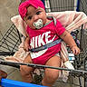 baby, infant, child, shopping_cart, supermarket, pink_bow, pacifier, nike_shirt, blanket, sitting, portrait, face, eyes, smiling, clothing, socks, grocery_store, metal_cart, indoor, shopping