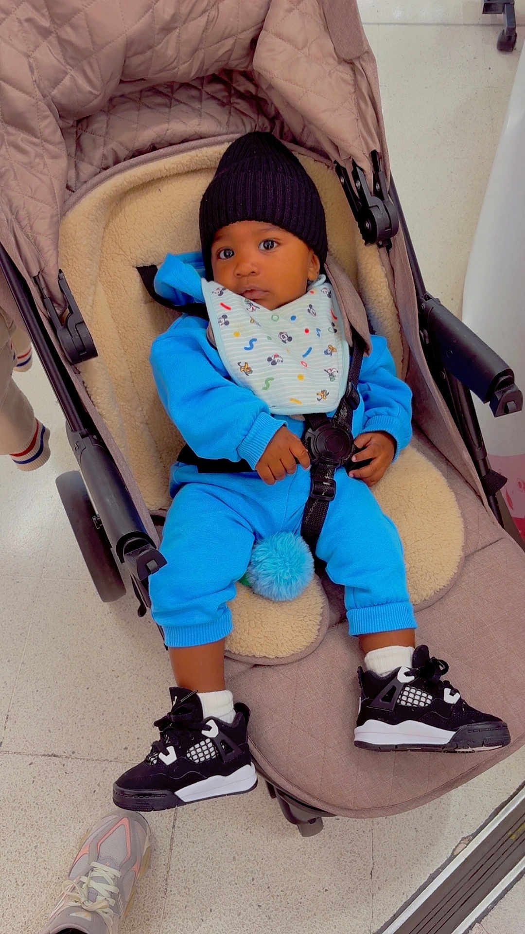 Kylo is registered to the contest to win money with this photo: toddler, baby, child, stroller, blue_outfit, beanie, sneakers, black_shoes, cushion, floor, indoor, curious, seated, person, cute, clothing, footwear, accessory, infant, strap