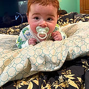 Lucius is registered to the contest to win money with this photo: baby, pacifier, bed, blanket, pattern, face, indoor, child, person, curious, sleepwear, furniture, television, clock, room, green_wall, portrait, closeup, hands, lying_down