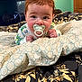 baby, pacifier, bed, blanket, pattern, face, indoor, child, person, curious, sleepwear, furniture, television, clock, room, green_wall, portrait, closeup, hands, lying_down