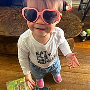 Mary is registered to the contest to win money with this photo: blue_jeans, casual_clothing, child, expression, floor, hair, heart_shaped, indoor, package, person, pink_shoes, playful, smiling, standing, sunglasses, toddler, toy_gun, white_shirt, wooden_floor, young_child