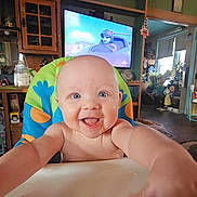 Parker joined the competition — help win amazing prizes! baby, child, smiling, high_chair, indoor, home, television, cartoon, furniture, person, happy, blue_eyes, bald, reaching, colorful, kitchen, living_room, smile, infant, cute