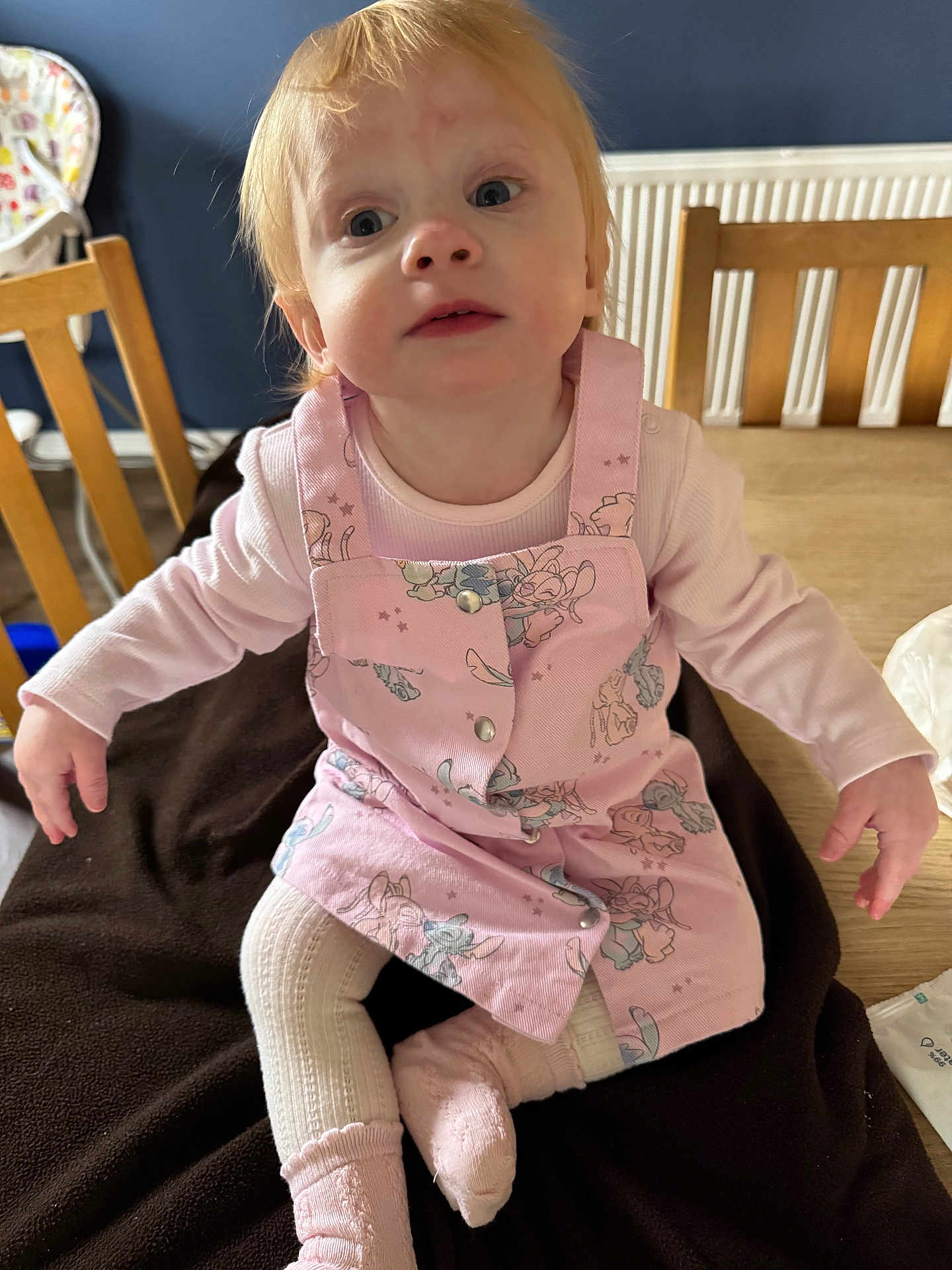 Mia-Louise joined the competition — help win amazing prizes! baby, blanket, child, cozy, curious_expression, face, floor, home, indoor, light_hair, long_sleeve, patterned_dress, person, pink_clothing, radiator, sitting, sock, table, toddler, wooden_chair