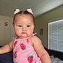 baby, child, pink_clothing, strawberry_pattern, hair_clips, indoor, bedroom, window_blinds, face, portrait, cute, chubby_cheeks, curly_hair, infant, person, sitting, soft_lighting, expression, young_child, casual_wear