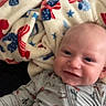 baby, smiling, onesie, blanket, patterned_clothing, indoor, infant, face, happy, cute, lying_down, cozy, soft_texture, child, portrait, closeup, person, comfortable, warm, sleepwear