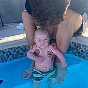 Kyle is registered to the contest to win money with this photo: baby, adult, pool, water, swim_trunks, child, person, curly_hair, skin, supporting_hands, outdoor, tiles, blue_water, swimming, summer, sunlight, family, parent, crouching, serious_expression