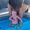 baby, adult, pool, water, swim_trunks, child, person, curly_hair, skin, supporting_hands, outdoor, tiles, blue_water, swimming, summer, sunlight, family, parent, crouching, serious_expression