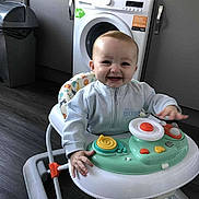 Cobi-Jai is registered to the contest to win money with this photo: baby, walker, smiling, kitchen, washing_machine, trash_bin, floor, clothing, child, person, toy, happy, indoor, appliance, cabinet, furniture, head, face, hand, playing