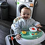baby, walker, smiling, kitchen, washing_machine, trash_bin, floor, clothing, child, person, toy, happy, indoor, appliance, cabinet, furniture, head, face, hand, playing