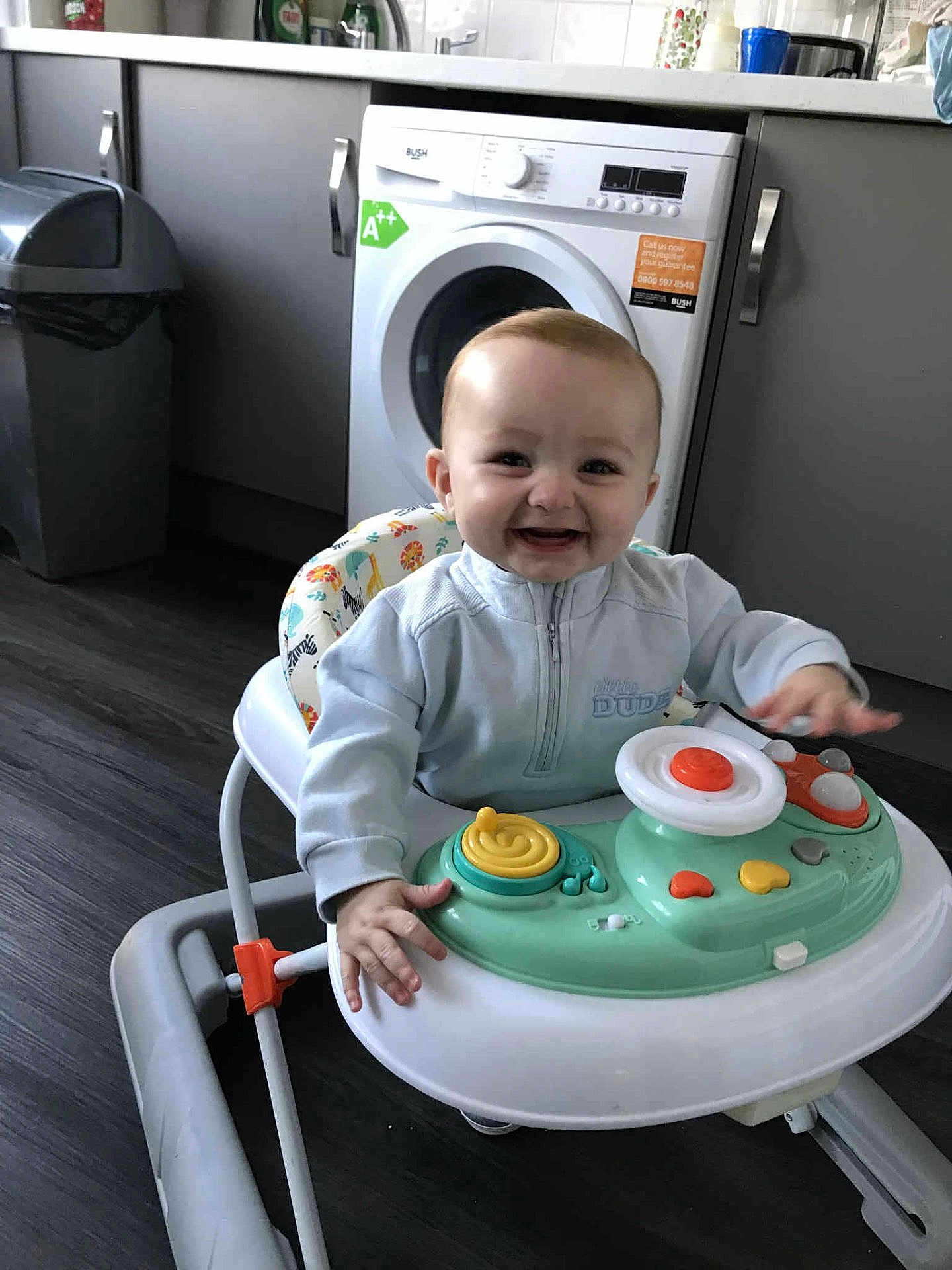 Cobi-Jai is registered to the contest to win money with this photo: baby, walker, smiling, kitchen, washing_machine, trash_bin, floor, clothing, child, person, toy, happy, indoor, appliance, cabinet, furniture, head, face, hand, playing