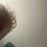 child, curly_hair, close_up, face_partial, blurry, indoor, wall, texture, colorful_object, corner, head, young, person, hair, background, light, shadow, skin, eyebrow, indoor_lighting