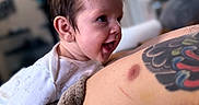 Atlas is registered to the contest to win money with this photo: baby, infant, child, smiling, laughing, portrait, close_up, indoor, candid, adult, parent, tattoo, arm, onesie, blanket, cuddling, happy, face, home, soft_lighting