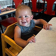 Rocco joined the competition — help win amazing prizes! toddler, child, high_chair, smiling, restaurant, table, wooden_chair, indoor, furniture, red_chair, person, casual_clothing, happy, young_child, dining, seat, hair, skin, face, arm