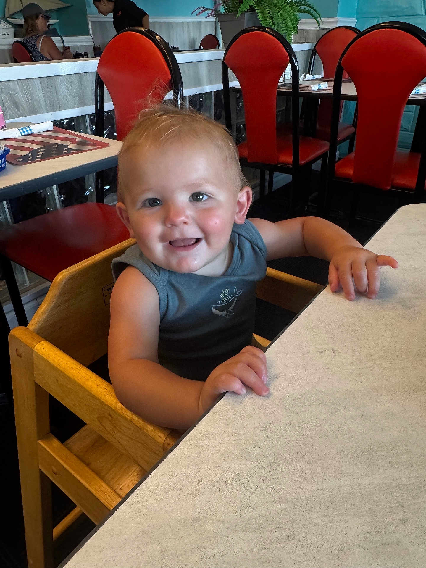 Rocco joined the competition — help win amazing prizes! toddler, child, high_chair, smiling, restaurant, table, wooden_chair, indoor, furniture, red_chair, person, casual_clothing, happy, young_child, dining, seat, hair, skin, face, arm