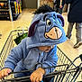 child, toddler, hooded_costume, eeyore, shopping_cart, supermarket, grocery_store, hands, hood, plush_design, person, adult_in_background, aisle, floor, bag, clothing, portrait, curious, shopping, trolley