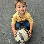Zahïm a rejoint le concours — aidez-le/la à gagner de superbes lots ! child, soccer_ball, asphalt, yellow_shirt, blue_jeans, smiling, happy, outdoor, playing, sport, young, boy, casual_clothing, holding, looking_up, hands, short_hair, daylight, recreation, fun