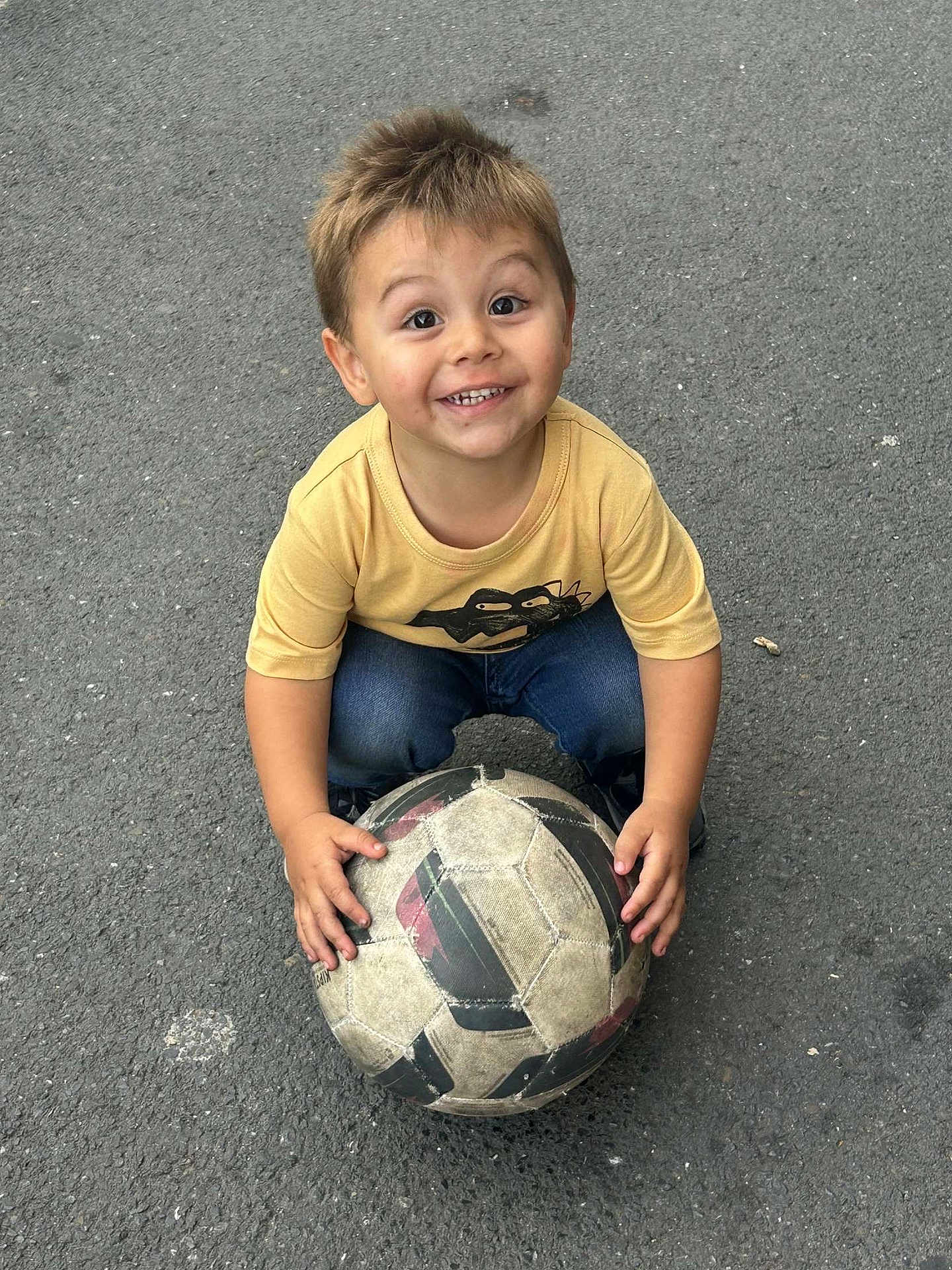 Zahïm a rejoint le concours — aidez-le/la à gagner de superbes lots ! child, soccer_ball, asphalt, yellow_shirt, blue_jeans, smiling, happy, outdoor, playing, sport, young, boy, casual_clothing, holding, looking_up, hands, short_hair, daylight, recreation, fun