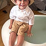 Zahïm a rejoint le concours — aidez-le/la à gagner de superbes lots ! toddler, child, hat, white_shirt, shorts, barefoot, smiling, sitting, indoor, furniture, modern_chair, happy, portrait, person, cute, young_child, floor, wooden_floor, casual_clothing, bright