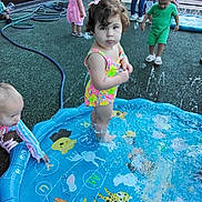 Angela joined the competition — help win amazing prizes! child, toddler, playground, water, splash_pad, pool, outdoor, summer, swimsuit, play, kids, fun, water_spray, daylight, curly_hair, colorful, toy, fence, grass, footwear