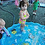 child, toddler, playground, water, splash_pad, pool, outdoor, summer, swimsuit, play, kids, fun, water_spray, daylight, curly_hair, colorful, toy, fence, grass, footwear