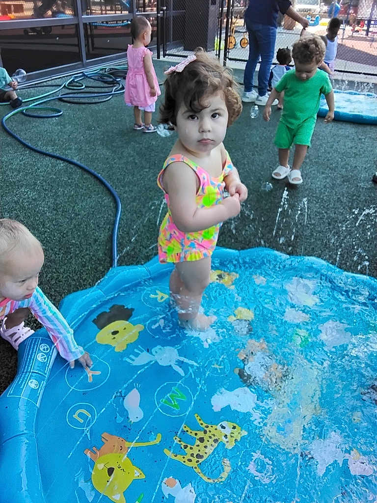 Angela joined the competition — help win amazing prizes! child, toddler, playground, water, splash_pad, pool, outdoor, summer, swimsuit, play, kids, fun, water_spray, daylight, curly_hair, colorful, toy, fence, grass, footwear