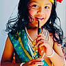 child, portrait, smiling, traditional_costume, crown, jewelry, flute, musical_instrument, red_flower, long_hair, makeup, cultural_attire, blue_clothing, yellow_skirt, pearl_necklace, bracelets, indoor, seated, colorful, photography