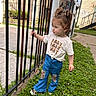 casual_clothing, child, concrete, curious, daylight, expression, fence, grass, hair_tied, jeans, leopard_print, nature, outdoor, person, residential, shirt, side_view, standing, toddler, young_child