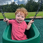 Theo is registered to the contest to win money with this photo: child, toddler, swing, playground, green_swing, curly_hair, red_hair, smiling, happy, outdoor, park, grass, trees, sky, clouds, pink_shirt, play, fun, person, daylight