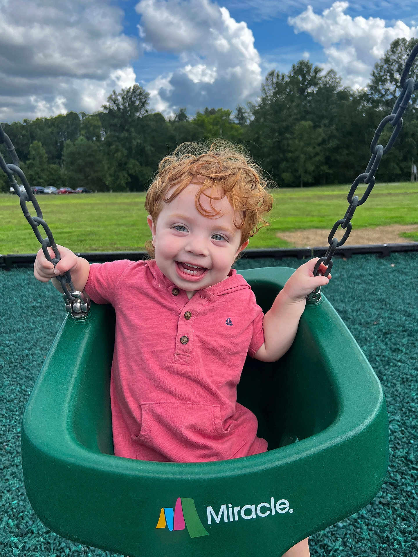 Theo is registered to the contest to win money with this photo: child, toddler, swing, playground, green_swing, curly_hair, red_hair, smiling, happy, outdoor, park, grass, trees, sky, clouds, pink_shirt, play, fun, person, daylight