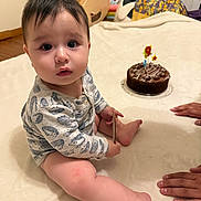 Jacob is registered to the contest to win money with this photo: baby, child, indoor, birthday_cake, candle, hands, blanket, onesie, toy, floor, wooden_toy, celebration, chocolate_cake, face, person, sitting, curious, cute, infant, home