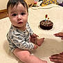 baby, child, indoor, birthday_cake, candle, hands, blanket, onesie, toy, floor, wooden_toy, celebration, chocolate_cake, face, person, sitting, curious, cute, infant, home