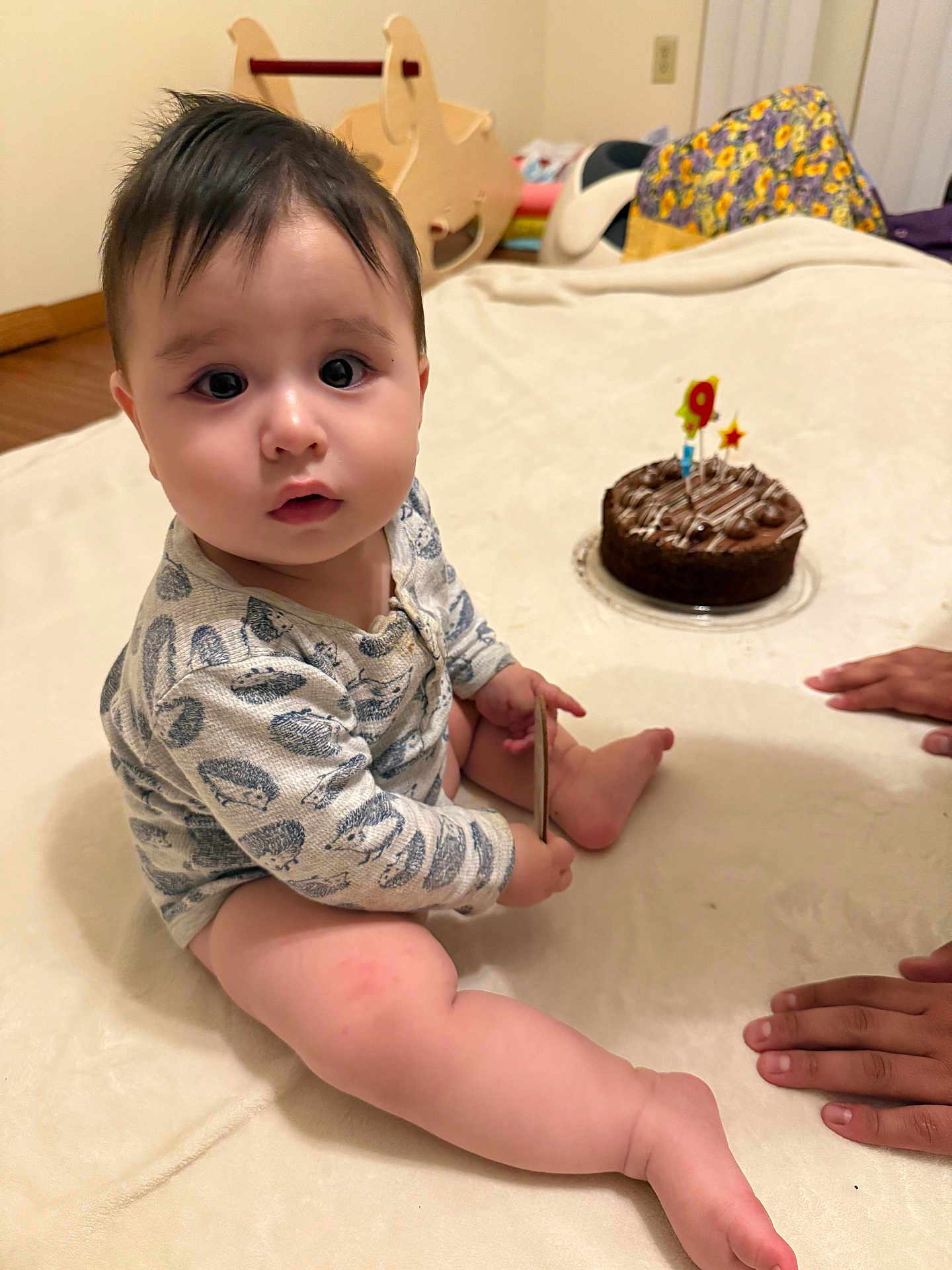 Jacob is registered to the contest to win money with this photo: baby, child, indoor, birthday_cake, candle, hands, blanket, onesie, toy, floor, wooden_toy, celebration, chocolate_cake, face, person, sitting, curious, cute, infant, home