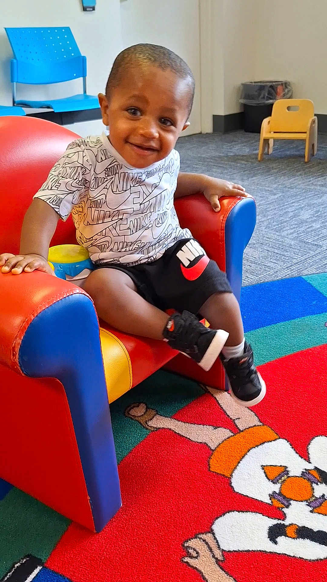 Antonio is registered to the contest to win money with this photo: toddler, child, smiling, indoor, armchair, colorful, nike, sneakers, carpet, playroom, furniture, child_furniture, happy, person, shorts, tshirt, play_area, floor, toy, room