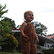 Layken is registered to the contest to win money with this photo: toddler, child, smiling, overalls, grass, outdoor, house, greenery, bushes, daylight, portrait, happy, person, nature, young_child, casual_clothing, front_yard, curly_hair, sunlight, playful
