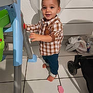 Naël participe au concours pour gagner de l'argent avec cette photo : toddler, child, barefoot, plaid_shirt, blue_pants, indoor, tile_floor, plastic_chair, standing, curious, smiling, young_child, person, home, toy, footwear_absent, furniture, baby, casual_clothing, expression