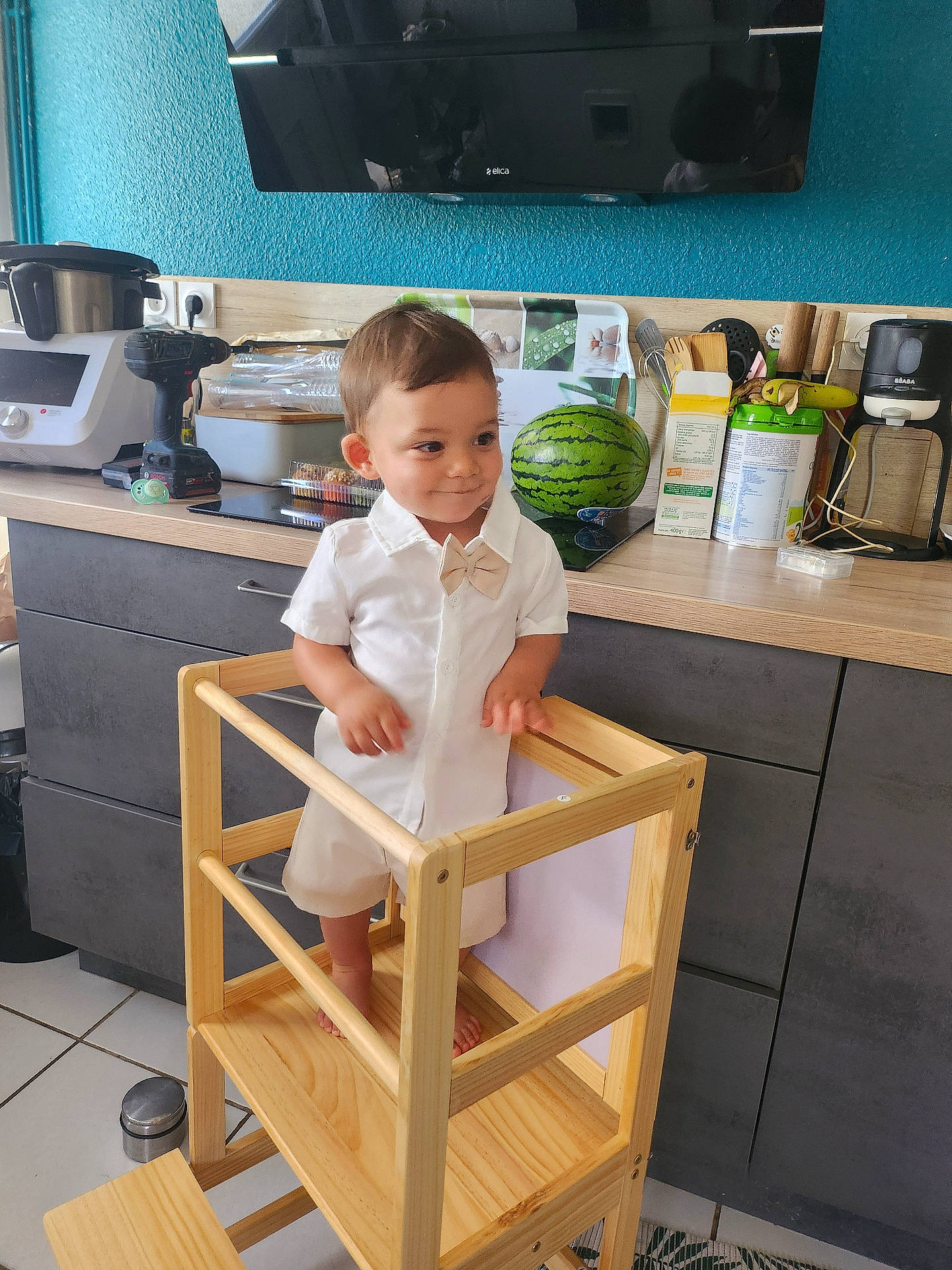 Naël participe au concours pour gagner de l'argent avec cette photo : appliance, barefoot, black_cabinet, bow_tie, child, cooking_tools, countertop, floor_tile, home, indoor, kitchen, kitchen_counter, modern_kitchen, person, smiling, toddler, utensils, watermelon, white_clothing, wooden_tower