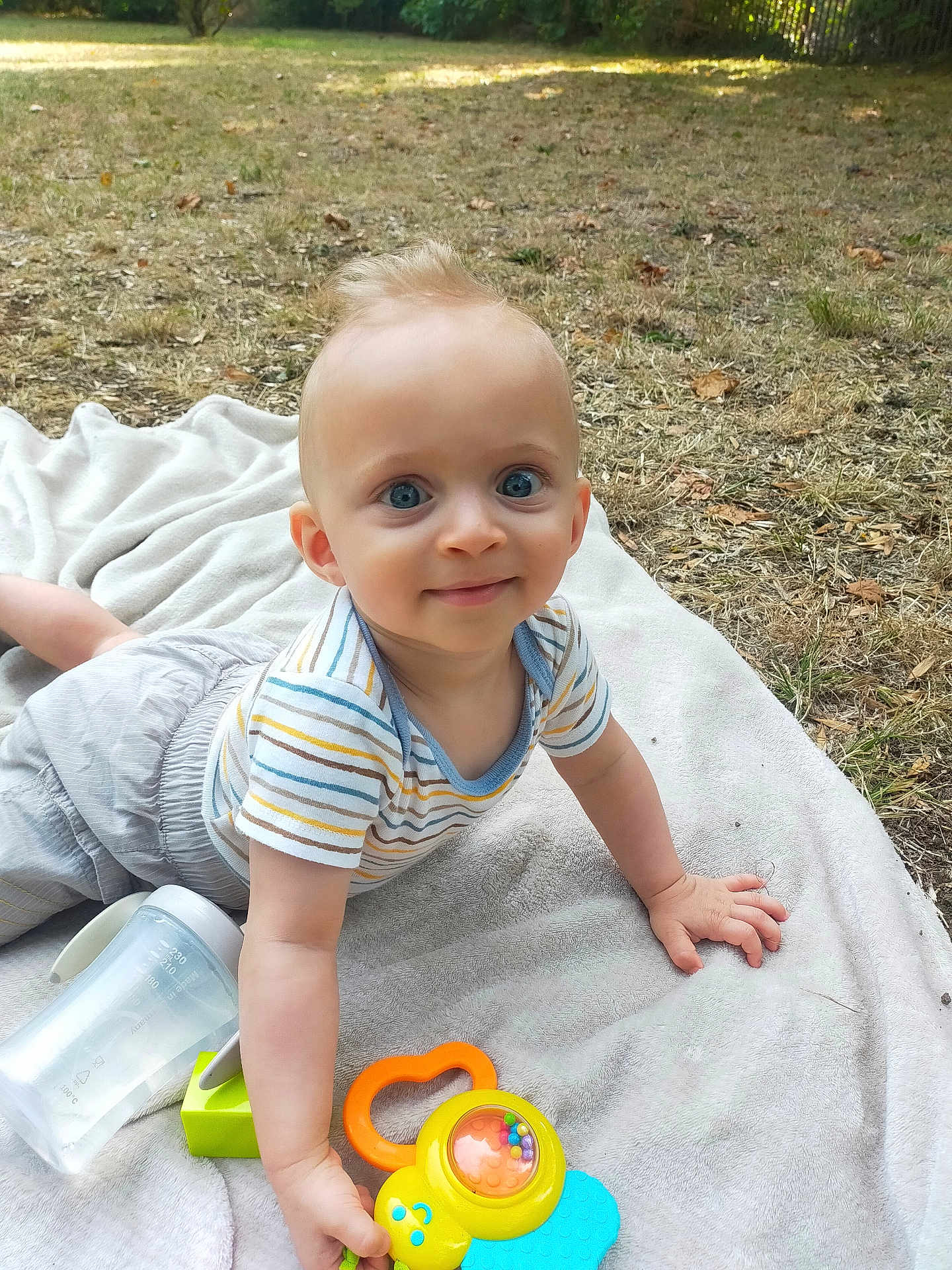 Marceau participe au concours pour gagner de l'argent avec cette photo : baby, child, toy, blanket, outdoor, grass, water_bottle, smile, blue_eyes, striped_shirt, shorts, hand, face, person, daylight, playing, cute, young, nature, happy