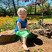 Jameson is registered to the contest to win money with this photo: child, boy, barefoot, rock, flowers, garden, outdoor, sunlight, greenery, tree, nature, summer, casual_clothing, grass, plant, daylight, young_child, sitting, bare_arms, curious_expression