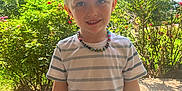 Jameson is registered to the contest to win money with this photo: child, boy, smiling, outdoor, sunlight, nature, plants, flowers, necklace, tshirt, casual_clothing, portrait, greenery, daytime, happy, standing, person, young, cute, garden