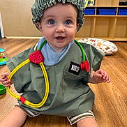 James is registered to the contest to win money with this photo: baby, child, infant, doctor_costume, nurse_costume, stethoscope, scrubs, shower_cap, smiling, happy, portrait, sitting, floor, indoor, classroom, wood_floor, toy, medical_play, shelving, nursery