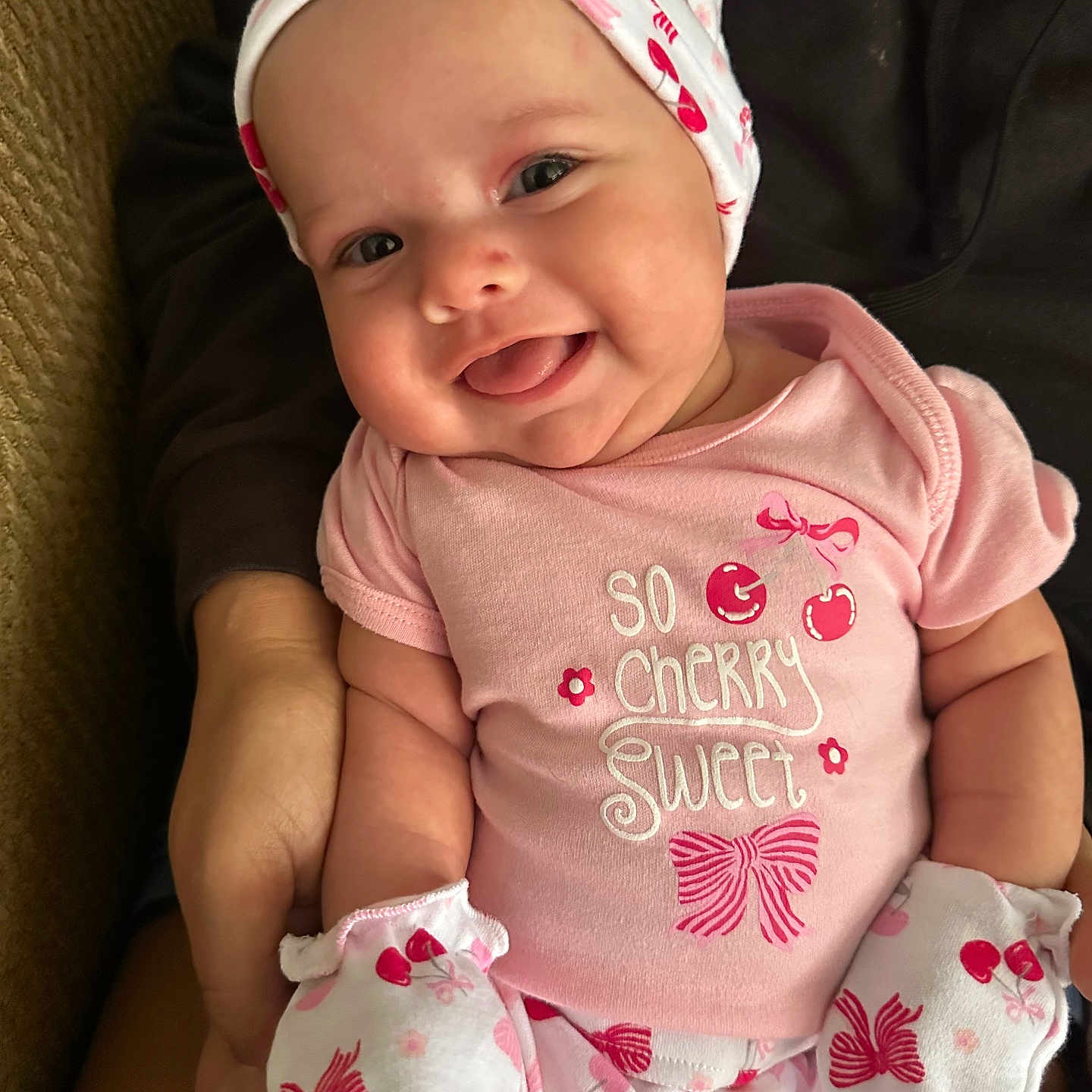Hailey joined the competition — help win amazing prizes! apparel, baby, cherry_pattern, child, closeup, cozy, cute, face, hand, happy, headwear, holding, indoor, infant, mittens, person, pink_clothing, sitting, smile, tongue_out