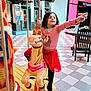 child, girl, carousel, horse, indoor, mall, pointing, red_skirt, striped_shirt, unicorn, happy, black_boots, floor_tiles, chair, storefront, window, smiling, toy, playful, person