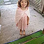child, girl, barefoot, pink_dress, cape, tongue_out, smiling, concrete_floor, green_mat, outdoor, wooden_lattice, playful, cute, standing, young, person, daylight, grass, casual, fun